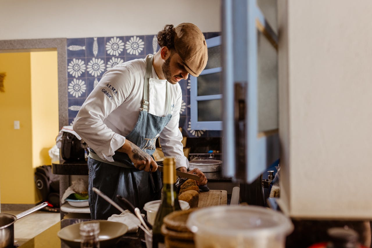 Chef wearing uniform and cap slicing bread in a contemporary kitchen setting.