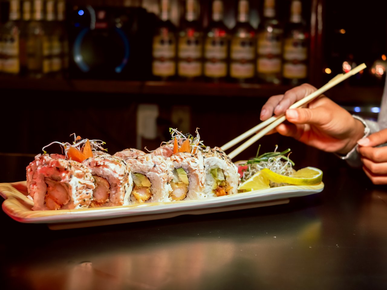 team-01 Close-up of gourmet sushi rolls with chopsticks in a cozy bar setting, highlighting fresh ingredients.