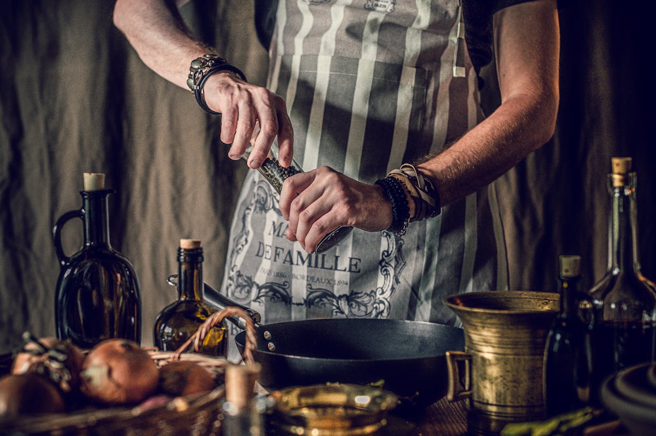 team-02 Crop faceless man in apron adding pepper in pan while cooking traditional dish standing at table with assorted ingredient and oil bottles in rustic kitchen